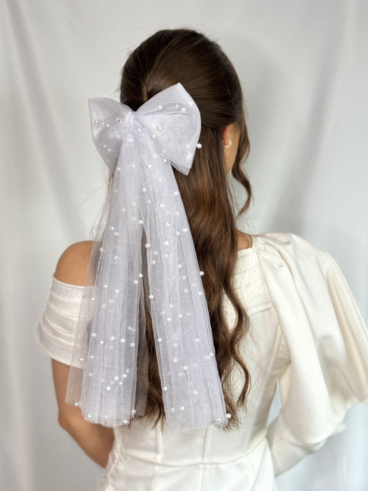 A woman wearing a white bridal hair bow with tulle and pearl embellishments attached to a metal clip, viewed from the back.